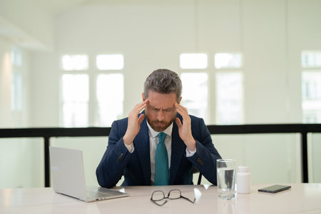 Millennial Businessman in office having headache. Business man in suit work on laptop at home office take off glasses suffer from migraine or headache. Tired exhausted man suffering from headache.の写真素材