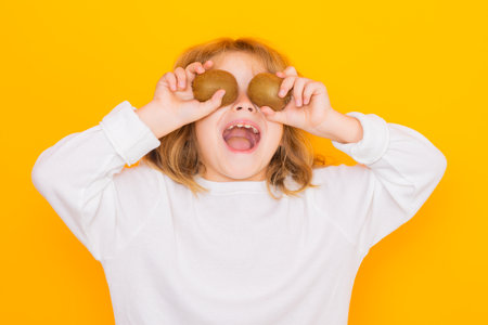 Kids face with fruits. Child hold kiwi in studio. Kiwi fruit. Studio portrait of cute kid boy with kiwi isolated on yellow.の写真素材