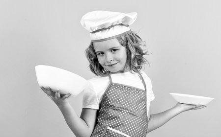 Funny kid chef cook with kitchen plate, studio portrait. Kid chef cook, studio portrait. Children cooking. Kid boy with apron and chef hat.の写真素材