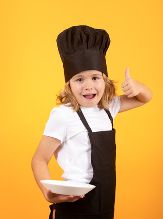 Funny child cook with cooking plate. Kid in cooker uniform and chef hat preparing food on studio color background. Cooking, culinary and kids food concept.の写真素材