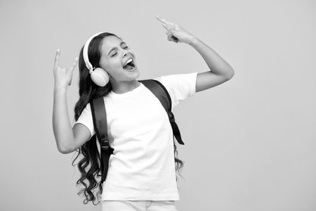 School girl in headphones with backpack ready to learn. School children with school bag on isolated yellow studio background.の写真素材