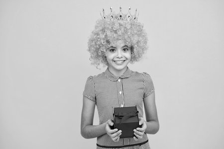 Funny princess in wig and crown. Teenager child girl holding present box isolated over yellow studio background. Present, greeting and gifting concept. Birthday holiday concept.の写真素材