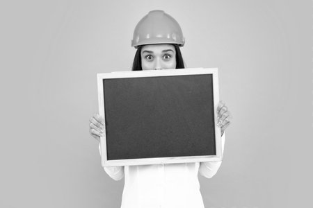 Happy woman holding and peaking over blank boar sign isolated on gray background. Smiling woman builder in helmet holding board for advertising sign. Sign board with copy space.の写真素材