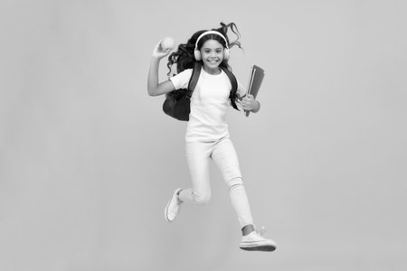 Back to school. Teenager school girl with backpack and headphones hold books ready to learn. School children on isolated yellow studio background.の写真素材