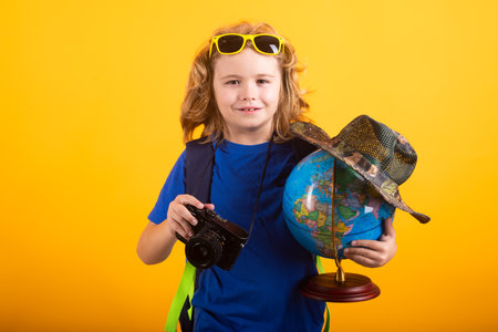 Cute blond kid wearing explorer hat and backpack on studio. Child explorer and adventure concept. Kid tourists hold world globe isolated on yellow background.の写真素材