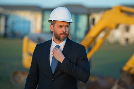 Construction business owner. Man in suit and hardhat halmet at building construction site. Civil engineer worker in front of house near excavator. Architect, supervisor investor, construction manager.の写真素材