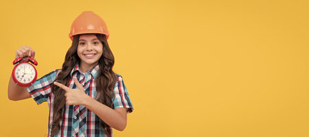 happy teen girl with curly hair in construction helmet pointing finger on alarm clock, in time. Child builder in helmet horizontal poster design. Banner header, copy space.の写真素材