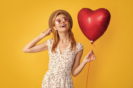 cheerful girl in straw hat and sunglasses hold love heart balloon on yellow backgroundの写真素材