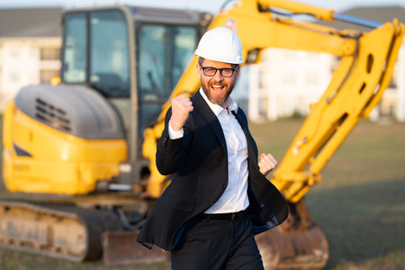 Construction manager in suit and helmet at a construction site. Construction manager worker or supervisor wearing hardhat in front of house. Supervisor construction manager near excavator. Renovation.の写真素材