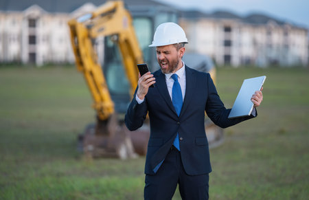 Buider man in suit and hardhat. Construction investor. Business man investor in front of construction site. Successful investor. Handsome man in suit and hardhat at building construction.の写真素材
