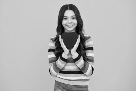 Cheerful lovely romantic teen girl hold red heart symbol of love for valentines day isolated on yellow background. Happy teenager, positive and smiling emotions of teen girl.の写真素材
