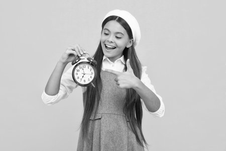 Teen student girl hold clock isolated on yellow background. Time to school. Teenager child with alarm clock showing time, late awakening. Happy girl face, positive and smiling emotions.の写真素材