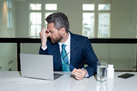 Stressed business man having headache at office. Tired businessman is working overtime and has headache. Overworked man with laptop at workplace, suffers from headache. Migraine from overwork.の写真素材