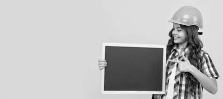 happy teen girl in helmet pointing finger on school blackboard with advertisement. Child builder in...