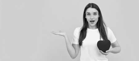amazed lady in white shirt with heart presenting product on yellow background, copy space, donation. Woman isolated face portrait, banner with copy space.の写真素材