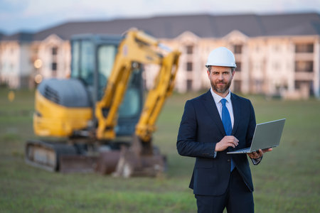 Architect at a construction site. Architect man in helmet and suit at modern home building construction. Architect with a safety vest and suit. Confident architect standing at house background.の写真素材