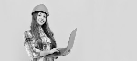 happy teen girl in helmet and checkered shirt using laptop, education online. Child builder in helmet horizontal poster design. Banner header, copy space.の写真素材