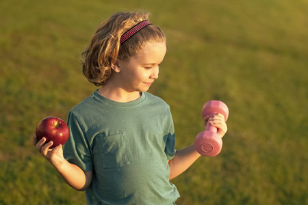 Healthy sporty child with apple and dumbbell outdoor in park. Cute child boy pumping up arm muscles with dumbbell. Fitness kids with dumbbells.の写真素材