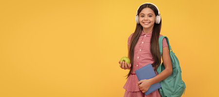 smiling school child in headphones carry backpack and workbook with apple for lunch. Portrait of schoolgirl student, studio banner header. School child face, copyspace.の写真素材