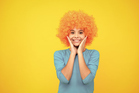 Birthday kids party. Funny kid in curly clown wig isolated on yellow background. Happy teenager portrait. Smiling girl.の写真素材