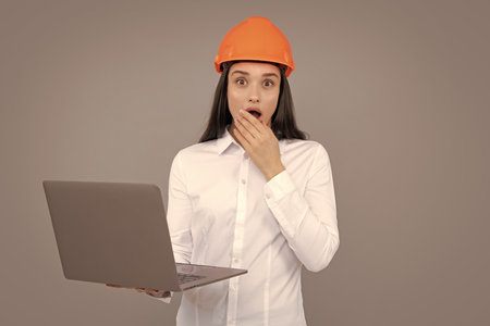Surprised shocked excited business woman with laptop computer, portrait on gray. Young woman in helmet hold laptop computer on gray background. Architect businesswoman.の写真素材