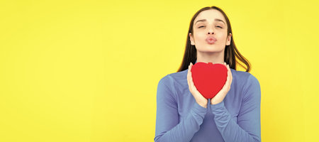 Beautiful girl holding valentines gift on yellow background. Portrait of young woman holding red paper heart. Woman isolated face portrait, banner with copy space.の写真素材