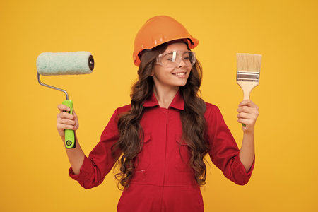 Child builder in helmet. Teenage girl painter with painting brush tool or paint roller. Child on repairing work isolated on yellow background. Kids renovation concept. Happy teenager girl.の写真素材