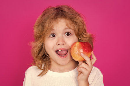 Kids face with fruits. Kid with apple in studio. Studio portrait of cute child hold apple isolated on pink background.の写真素材