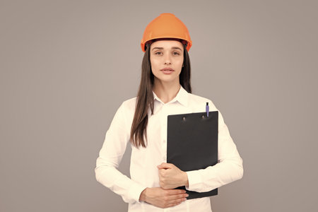 Serious woman worker in protective helmet with clipboard isolated on grey background. Young woman construction manager. Architect woman, female worker in hardhat helmet.の写真素材