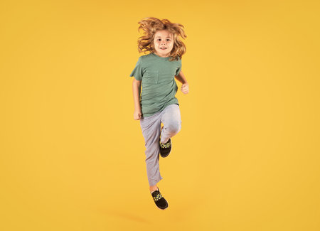 Energetic kid boy jumping and raising hands up on isolated studio background. Full length body size photo of jumping high child boy, hurrying up running fast on yellow background.の写真素材