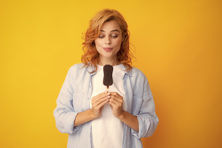 Young woman eats delicious ice cream, enjoys frozen dessert, dressed in casual clothes, isolated on yellow background.の写真素材