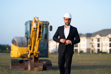 Construction manager in suit and helmet at a construction site. Construction manager worker or supervisor wearing hardhat in front of house. Supervisor construction manager near excavator. Renovation.の写真素材