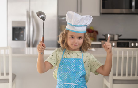 Fun kids kitchen. Funny little kid chef cook wearing uniform cook cap and apron cooked food in the kitchen. Kids are preparing food, bake cookies in the kitchen.の写真素材