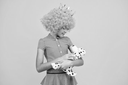 Teenager princess child in fancy clown wig hug toy. Teen child in queen crown isolated on yellow background. Princess girl in tiara. Prom party.の写真素材
