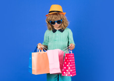 Child boy with many shopping bags in studio. Funny little kid in summer fashion kids clothes with shopping bags on isolated blue background.の写真素材