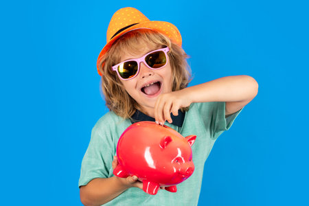 Saving money for travel. Portrait of a little boy putting money on a moneybox. Child saving money in a piggybank on blue background.の写真素材
