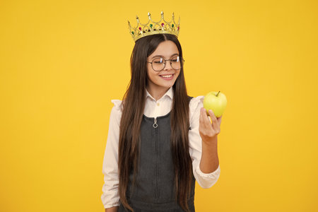 Princess child in queen crown hold apple isolated on yellow background. Teenage girl wear diadem. Happy girl face, positive and smiling emotions.の写真素材
