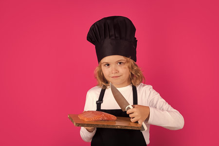 Kid cook hold cutting board with fish salmon steak and knife. Chef child preparing healthy food. Studio portrait of child with chef hats.の写真素材