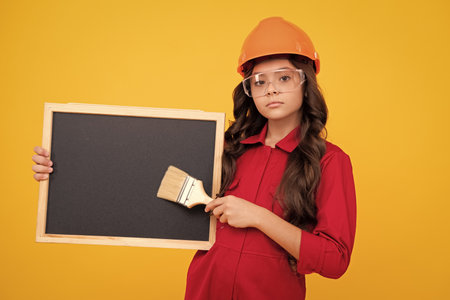 Child builder in helmet. Teenage girl painter with painting brush tool or paint roller. Child on repairing work isolated on yellow background. Kids renovation concept. Blackboard for copy space.の写真素材