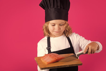 Kid cook hold cutting board with fish salmon steak and knife. Kid in cooker uniform and chef hat preparing food on studio color background. Cooking, culinary and kids food concept.の写真素材