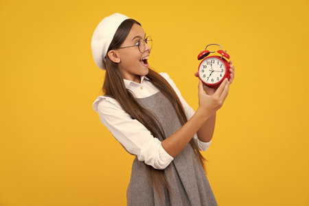 Teenager child hold clock isolated on yellow studio background. Teenager child with alarm clock showing time. Excited face, cheerful emotions of teenager girl.の写真素材