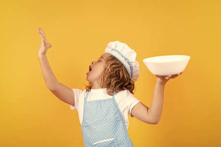 Funny kid chef cook with kitchen plate, studio portrait. Kid in cooker uniform and chef hat preparing food on studio color background. Cooking, culinary and kids food concept.の写真素材