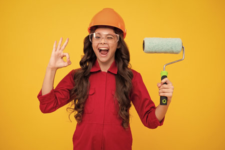 Excited teenager girl. Painter in workshop. Renovation and repair. Teen girl in helmet with painting brush. Child in hard hat. Kid builder painter on construction site.の写真素材