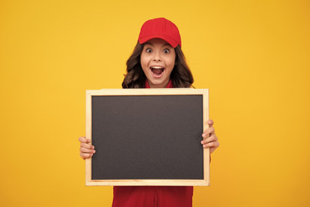 Excited teenager girl in red building uniform and cap. Teenage girl worker hold blackboard isolated on yellow background. Kids renovation concept. Copy space on empty board, mock up.の写真素材