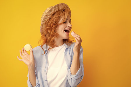 Cute young woman hold macaroon. Beautiful smiling young girl eating colorful macaroons over yellow background.の写真素材