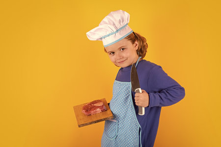 Kid cook hold cutting board with meat beef steak and knife. Cooking, culinary and kids. Little boy in chefs hat and apron on studio isolated background.の写真素材