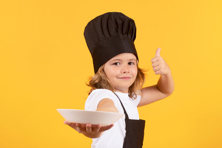 Funny child cook with cooking plate. Cooking children. Chef kid boy in form of cook. Child boy with apron and chef hat preparing a healthy meal on studio isolated background. Cooking process.の写真素材