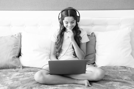 Teenage girl child in headphones relax on bed at home listen to music using laptop. Child in earphones browse internet on computer.の写真素材