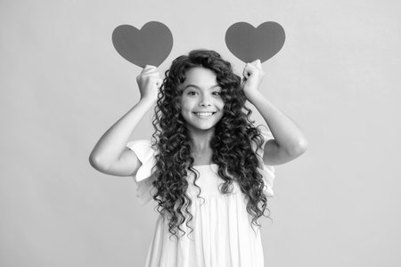 Happy teenager portrait. Cheerful lovely romantic teen girl hold red heart symbol of love for valentines day isolated on yellow background. Smiling girl.の写真素材