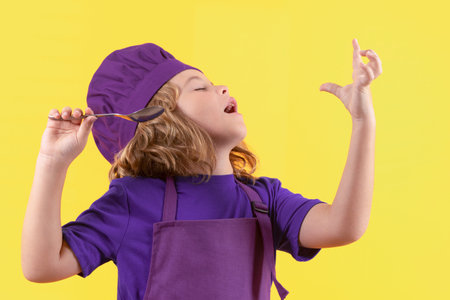 Funny kid chef cook with spoon, studio portrait. Child in cook uniform. Chef kid isolated on yellow background. Cute child to be a chef. Child dressed as a chef hat.の写真素材